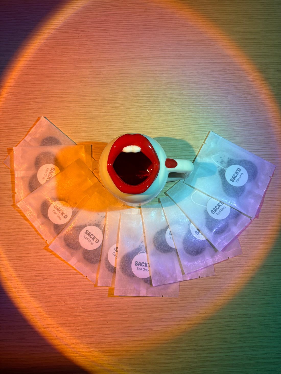 A red-lipped mug surrounded by individually wrapped SACK'D tea bags under colored light